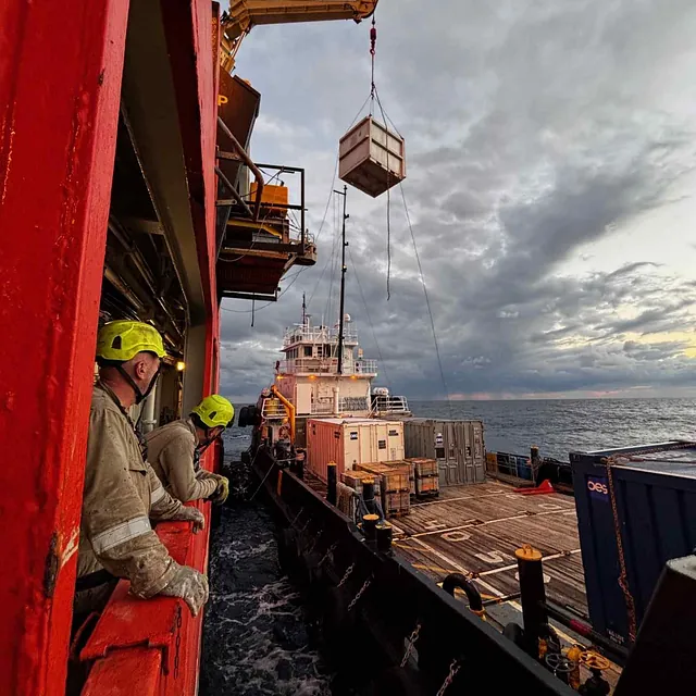 Workers in protective gear lifting a container between two ships with a crane, surrounded by ocean and an overcast sky.