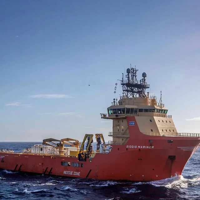 A supply ship in red sailing on the ocean, with equipment on deck and waves around the hull.